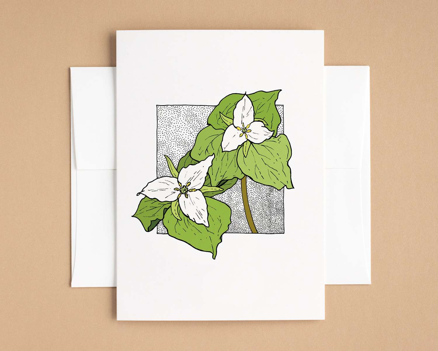 A vertical white card depicts two trillium flowers against a black stipple background. The card lies on top of a white envelope, which lies on top of a brown backdrop.
